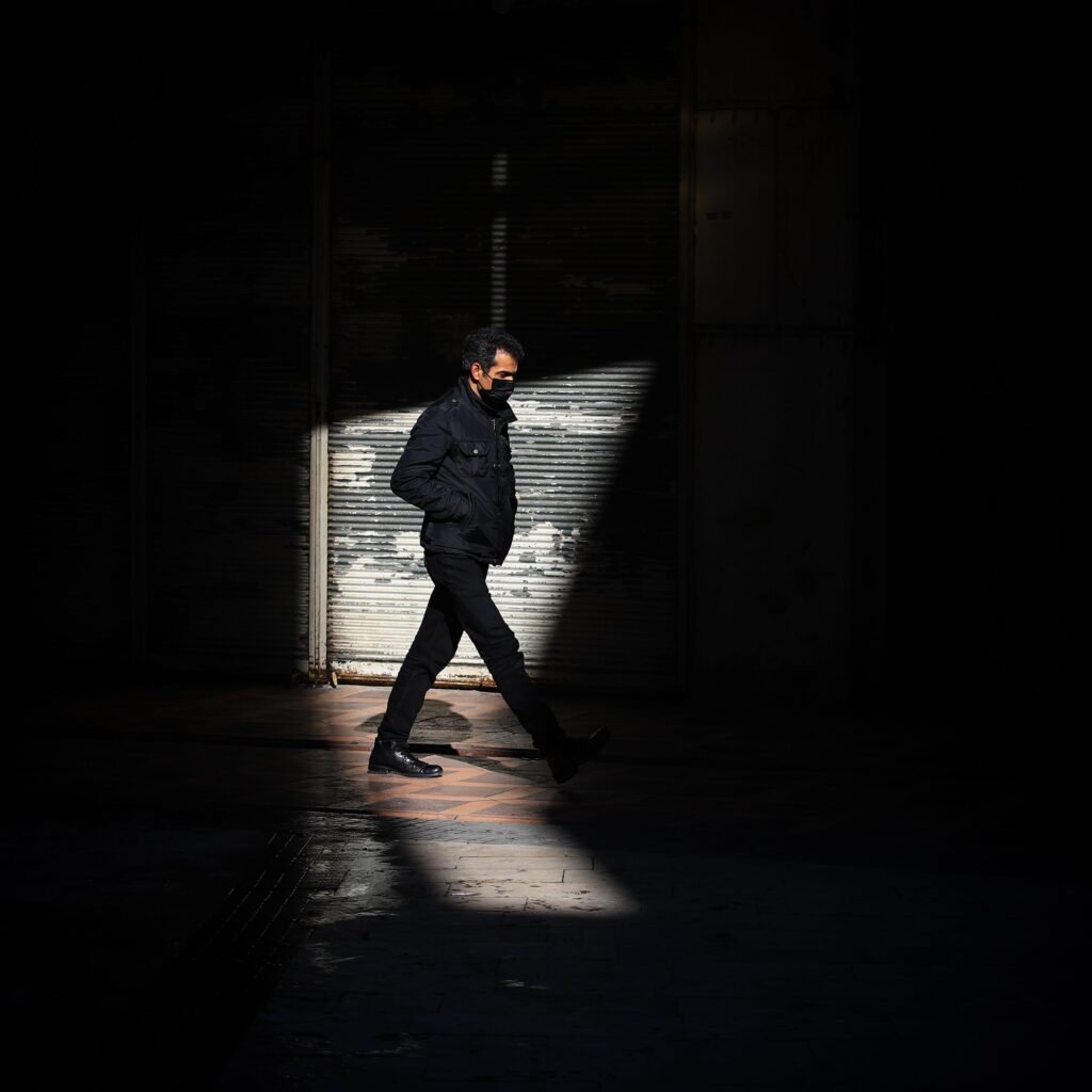Man in face mask walks through shaft of sunlight in urban setting.